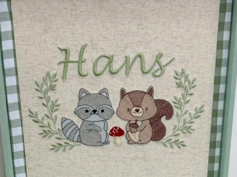 O nome Hans bordado em verde sobre um guaxinim de �culos e um esquilo segurando uma bolota, com um cogumelo vermelho entre eles e coroa de ramos