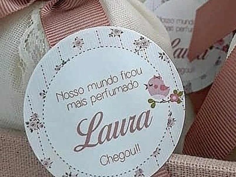 Tag redonda floral com o texto Nosso mundo ficou mais perfumado / Laura / Chegou, presa ao la�o rose com p�rola do sach� de cetim e renda