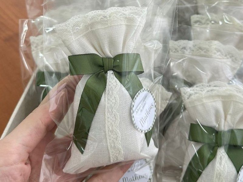Sach� de tecido branco com renda franzida no fecho e uma fita de renda descendo verticalmente pela frente, sob la�o de gorgur�o verde